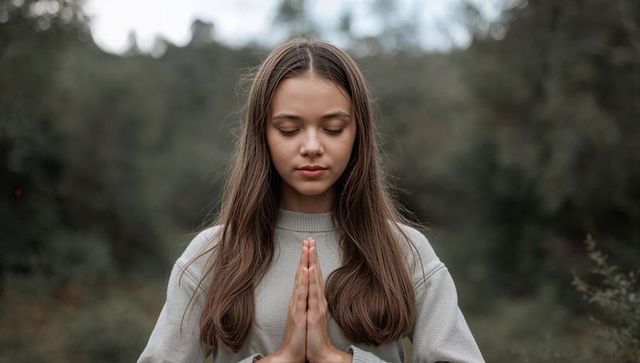 Teenage girl meditating with hands in prayer at forest edge, serene mindfulness portrait