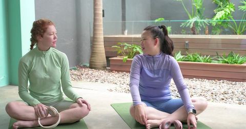 Diverse women sitting on yoga mats chatting after workout with pilates rings in tropical courtyard
