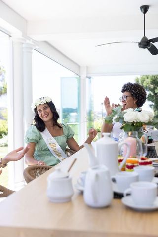Bridal brunch on sunlit veranda with bride-to-be wearing sash and floral crown laughing
