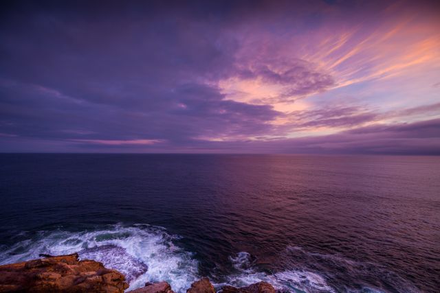 Dramatic sunset illuminating ocean with purples and pinks. Rocky shoreline in foreground. Ideal for use in travel promotions, environmental posters, or relaxation-themed content.