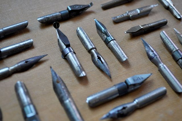 This image shows a variety of vintage metal calligraphy pen nibs arranged on a light background. Ideal for illustrating topics related to writing, calligraphy, and art supplies. Suitable for blogs about hand lettering, stationery shops, or history of writing implements. Can also be used in graphics for artistic and creative themes in advertising or educational materials.