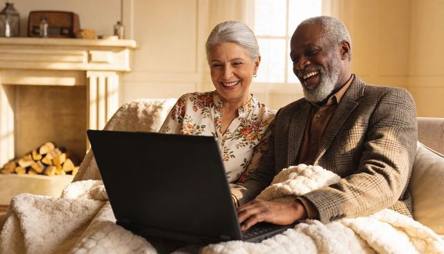 Laughing senior black man and white woman using laptop on cozy sofa by fireplace