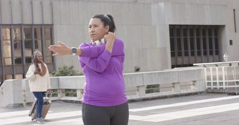 Diverse women stretching on city plaza with headphones and smartwatch, traveler checking phone