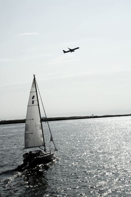 This image features a picturesque scene with a sailboat moving on the ocean water while an airplane flies overhead. The clear sky and sparkling water create a serene and charming atmosphere. Useful for themes related to travel, transportation, and freedom. Suitable for travel agencies, transportation services, and tourism campaigns.