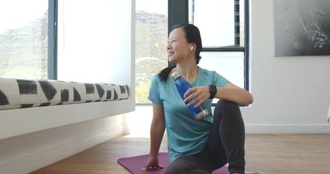Asian woman relaxing after yoga at home holding water bottle smiling by sunlit window
