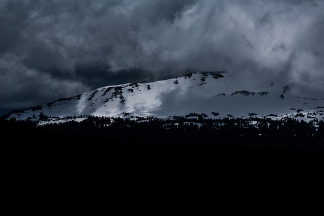 Image showcases a snow-capped mountain nestled under dark, stormy clouds. The dramatic lighting highlights the natural beauty of the rugged mountain and provides an atmospheric touch. Ideal for use in projects related to nature, adventure, outdoor activities, and environmental themes.