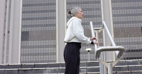 Mature woman standing on urban steps holding handrail beside office tower wearing hoodie