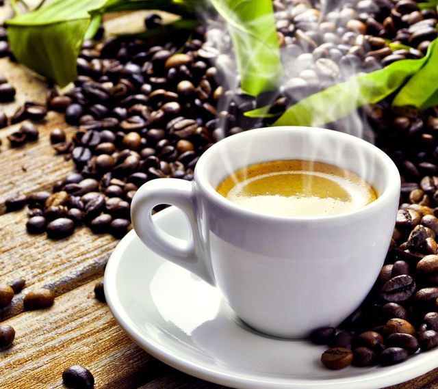 Steaming hot espresso in white cup surrounded by coffee beans and green leaves on wooden table. Ideal for coffee shop promotions, blog articles about coffee, and social media posts related to morning rituals and relaxation.