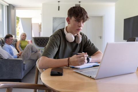 Teen studying on laptop with headphones while parents relaxing in modern living room