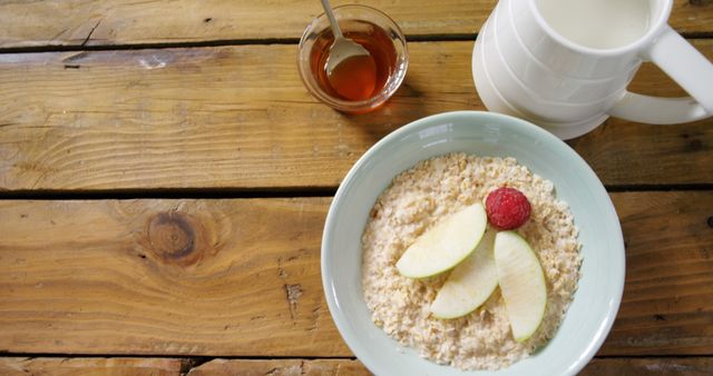 Oatmeal topped with apple slices and a raspberry sits in a light green bowl on a rustic wooden table. Milk in a jug and honey in a glass bowl with a spoon accompany the meal. Perfect for promoting healthy breakfast options, nutrition content, or rustic kitchen themes.