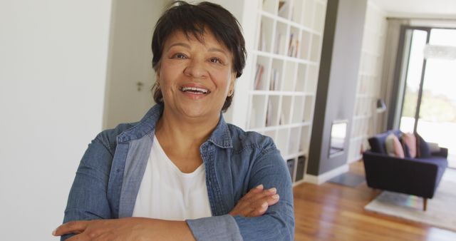 Middle-aged woman standing indoors with arms crossed, smiling confidently. She is in casual clothing, conveying relaxation and happiness. Ideal for advertising home decor, lifestyle products, and wellness services, or for illustrating articles about aging with confidence and happiness.
