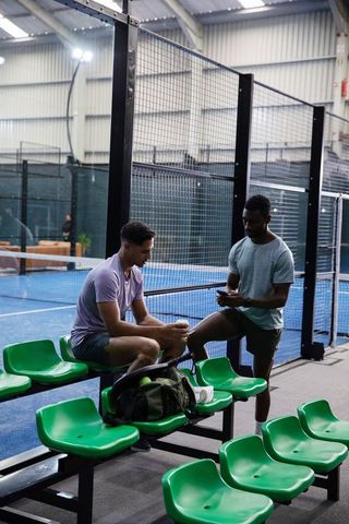 Two players chatting during break on indoor padel court with green spectator seats