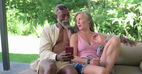 Mature diverse couple relaxing on sunlit patio sofa, sharing smartphone and laughing