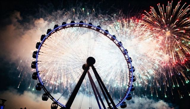 Illuminated ferris wheel silhouetted against explosive fireworks over nighttime waterfront