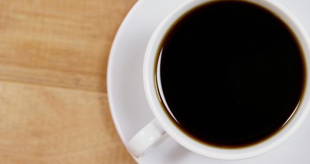 This image shows a top view of a cup of black coffee resting on a white saucer on a wooden table. Perfect for use in advertising coffee shops, morning routines, or promoting beverages. Ideal for articles related to coffee culture or as a background for quotes or social media posts about coffee.