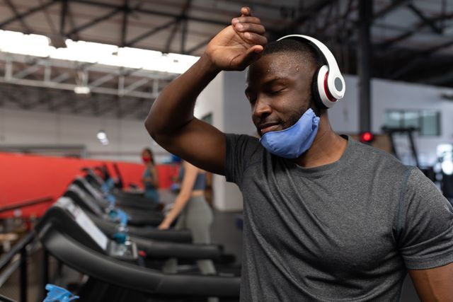 African American man wearing headphones and a facemask pulled down under his chin, wiping sweat from his forehead while at the gym. Ideal for use in articles or advertisements related to fitness, health, gym safety protocols, or active lifestyles.