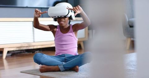 African american girl playing with vr headset at home, sitting cross-legged on rug