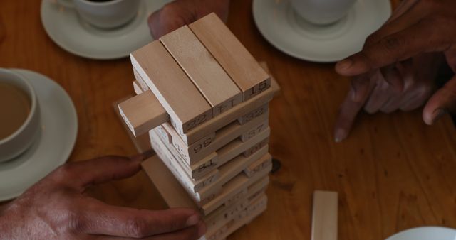 Hands are in motion while engaging in a Jenga game at a wooden table. Cups of coffee are visible in the background, indicating a relaxed and sociable atmosphere. Suitable for themes of family fun, teamwork, strategic games, and indoor activities.