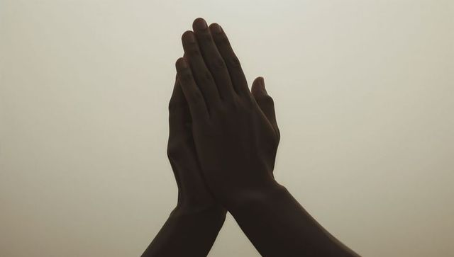 Dark-skinned hands pressed together in prayer gesture on soft beige gradient background