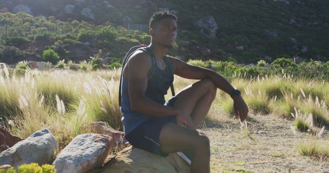 African american man exercising outdoors hiking resting on a rock in countryside on a mountain. fitness training and healthy outdoor lifestyle.