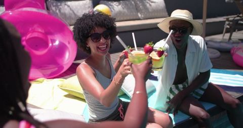 African american friends toasting tropical cocktails by poolside float laughing in sunlight