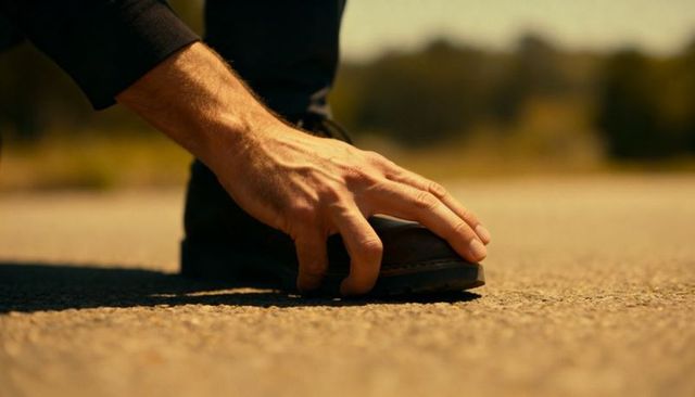 Male-presenting hand steadying brown leather shoe on sunlit asphalt closeup with texture