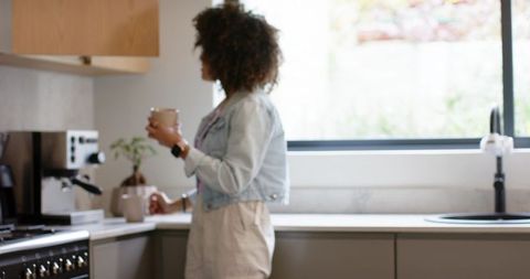 African american woman holding coffee mug at sunlit modern kitchen counter, relaxing