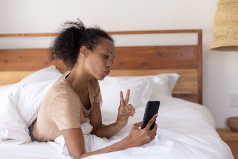 African american woman relaxing in bed video calling on smartphone, blowing kiss