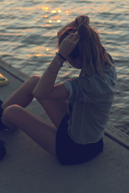 Young woman sitting calmly by the water during sunset, adjusting her hair. Peaceful and contemplative feel makes it ideal for themes of relaxation, self-reflection, and solitude. Suitable for promoting mental health, leisure activities, and lifestyle articles centered on unwinding or taking a moment for yourself.