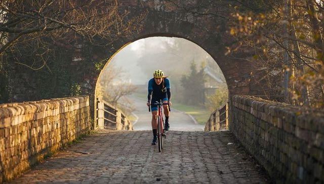 Cyclist riding under stone arch bridge, leaning and pedaling in yellow helmet and blue kit