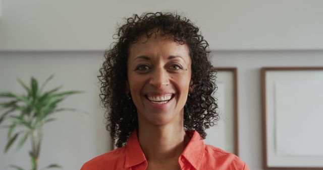 Smiling woman with curly hair dressed in an orange shirt posing indoors. Background has wall art and a plant, suggesting a home environment. Great for use in lifestyle, home decor, or self-confidence themes.