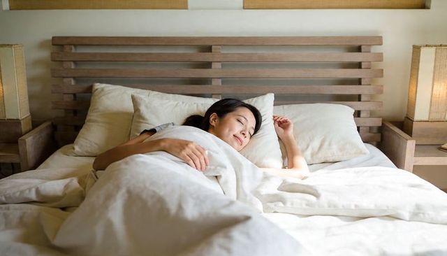 Asian woman sleeping peacefully on neutral king bed with white linens and wooden headboard