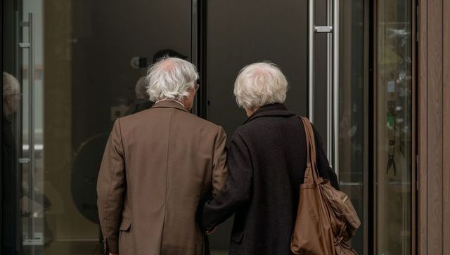Silver-haired couple walking through glass doors into urban lobby carrying satchel