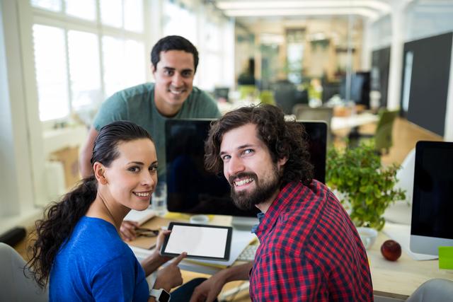 Group of graphic designers collaborating at a modern office workspace. One person is holding a digital tablet, and they are all smiling and looking at the camera. This image is perfect for illustrating teamwork, creative processes, collaboration in a professional environment, and modern office settings. Ideal for use in articles, blogs, websites, and marketing materials focused on teamwork, creativity, and office culture.