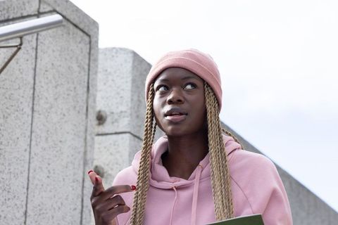 Young african american student pointing and holding folder, wearing pink beanie and hoodie