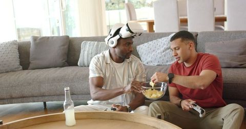 Diverse men sharing chips and gaming with controllers and VR headset in modern living room
