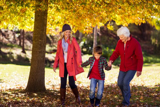 Boy walking hand in hand with mother and grandmother in a park during autumn. Leaves are falling, creating a colorful and vibrant atmosphere. Ideal for use in family-oriented content, advertisements for outdoor activities, or promotions for autumn events.