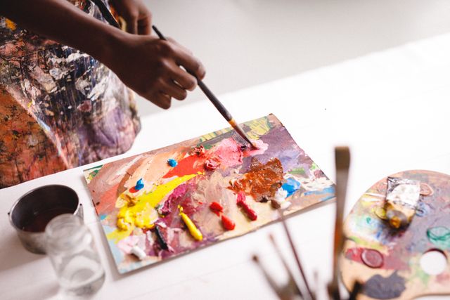 African American male artist mixes vibrant paints on a palette in an art studio environment. Useful for advertising art classes, creative workshops, and artistic inspiration. Suitable for illustrating concepts of creativity, artistic process, and hands-on painting techniques.