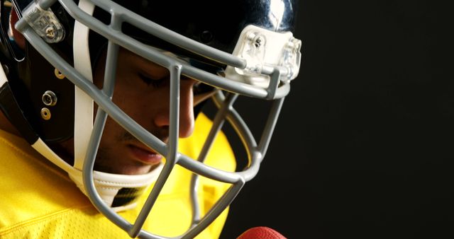Close-up shows football player wearing helmet holding ball against dark background. Useful for illustrating themes of sportsmanship, focus, determination, and team sports.