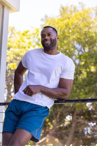 Smiling african american man leaning on balcony railing wearing white tee and blue shorts