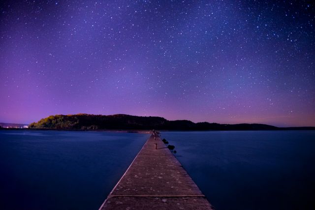 This image presents a long pier stretching out into a lake under a clear, star-filled night sky. The calm waters of the lake create a serene and tranquil atmosphere. Suitable for use in travel blogs, nature websites, or calming and inspirational backgrounds.