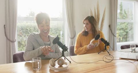 Two women recording podcast at home laughing and adjusting microphones and headphones
