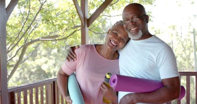 Elderly African American couple smiling while holding yoga mats outdoors, enjoying a sunny day on a wooden deck surrounded by trees. This image is perfect for promoting senior wellness, fitness programs, healthcare services, and active lifestyles. It can also be used in advertisements and articles about healthy aging, retirement, and outdoor activities for seniors.