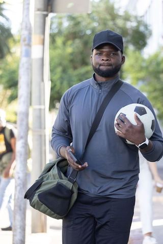 African man urban athlete holding soccer ball and phone walking to training with duffel