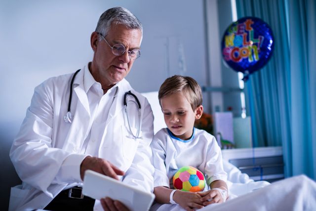 Doctor showing medical report to young patient using a digital tablet in a hospital room. The child is sitting on a hospital bed holding a colorful ball, with a 'Get Well Soon' balloon in the background. This image can be used for healthcare, pediatric care, medical technology, patient care, and hospital-related themes.