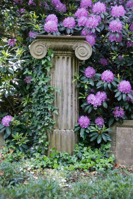 Ancient stone column standing amidst lush green foliage and vibrant purple flowers outdoors. Ivy climbing up the base, creating a picturesque scene. This image can be used for themes related to historical architecture, garden design, and nature's beauty.