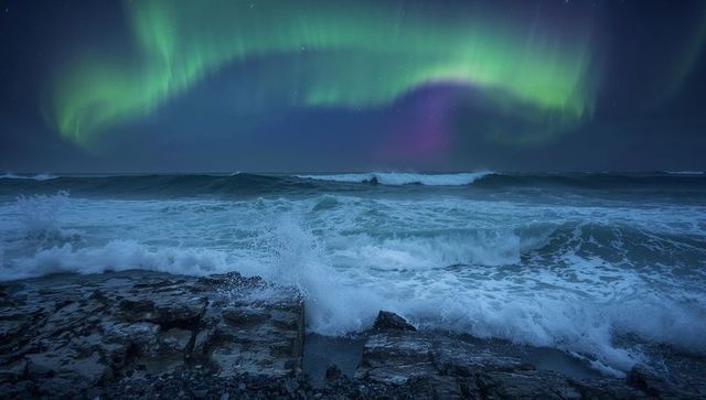 Aurora arcing over jagged rocky coast with crashing waves spraying sea foam