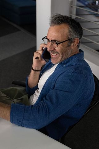 Smiling middle-aged man talking on smartphone at modern office desk, casual professional