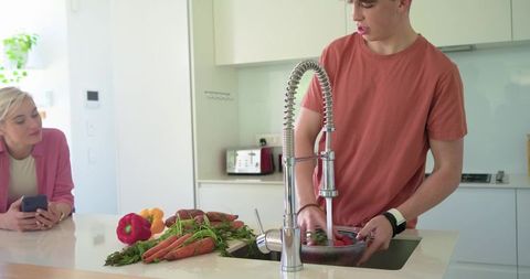 Woman watching teenage male rinsing fresh vegetables at modern kitchen island with coil faucet
