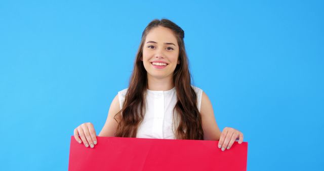 Young woman is holding a red board with a cheerful smile, standing against a vivid blue background. Ideal for use in advertising, promotions, marketing materials, or as a blank canvas for adding custom text or graphics.